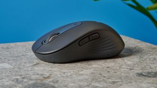 a black mouse with a scroll wheel, two side buttons, and grooved edges, with a logi logo, sits atop a pale table with a blue background